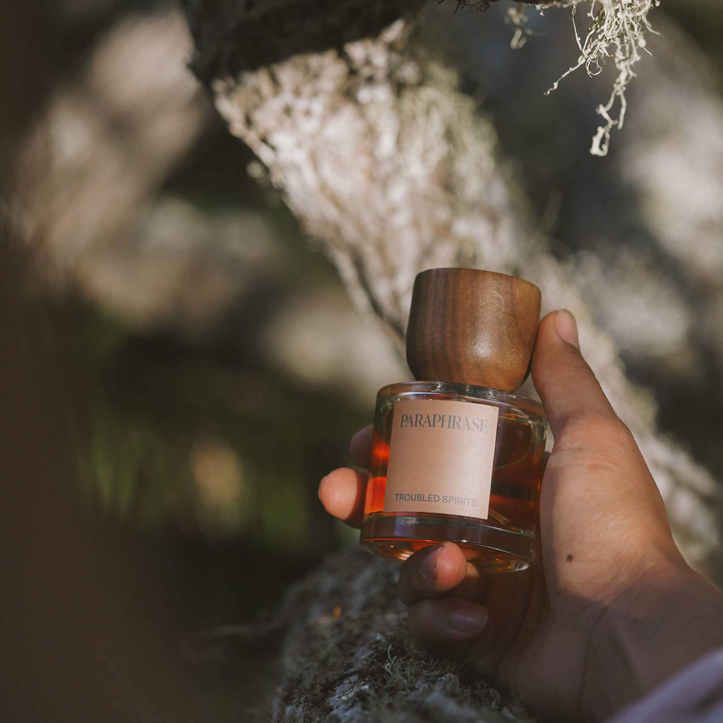 Hand holding Paraphrase Troubled Spirits amber perfume against mossy tree bark