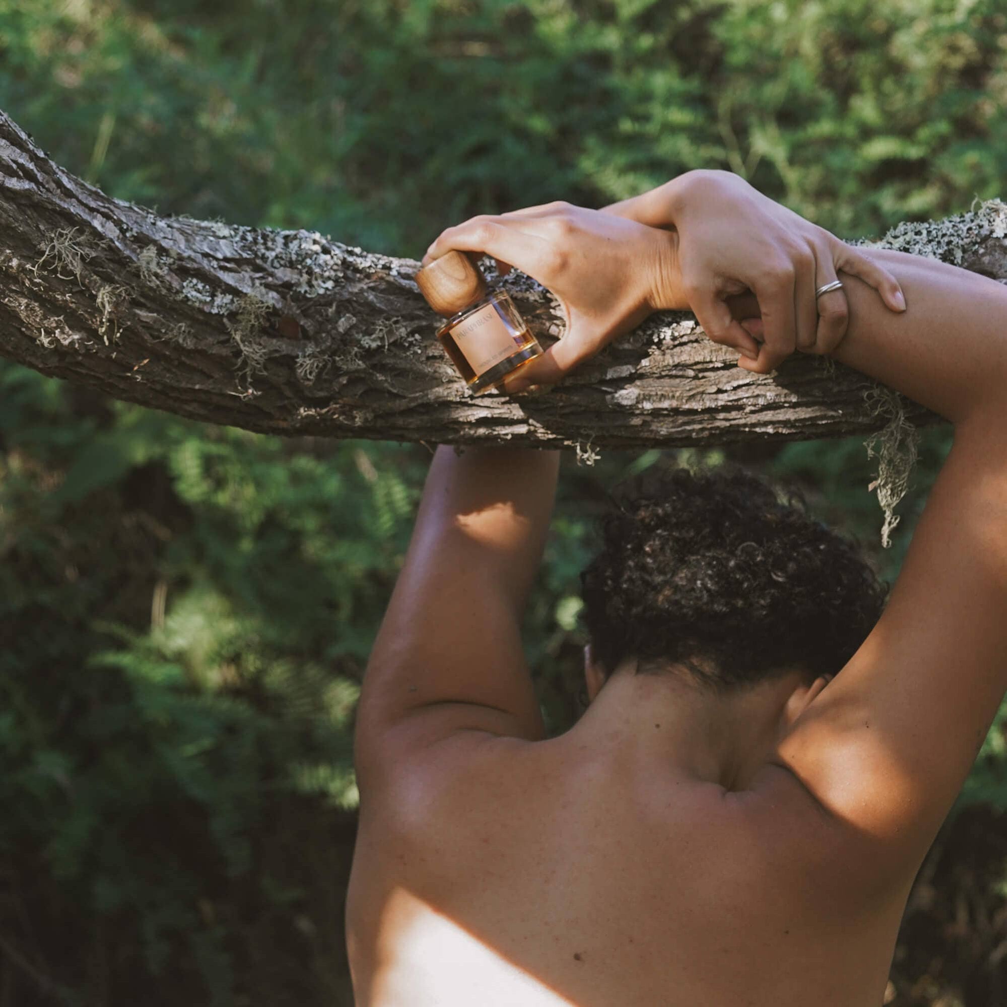 Shirtless person holding Paraphrase Troubled Spirits perfume bottle in forest setting
