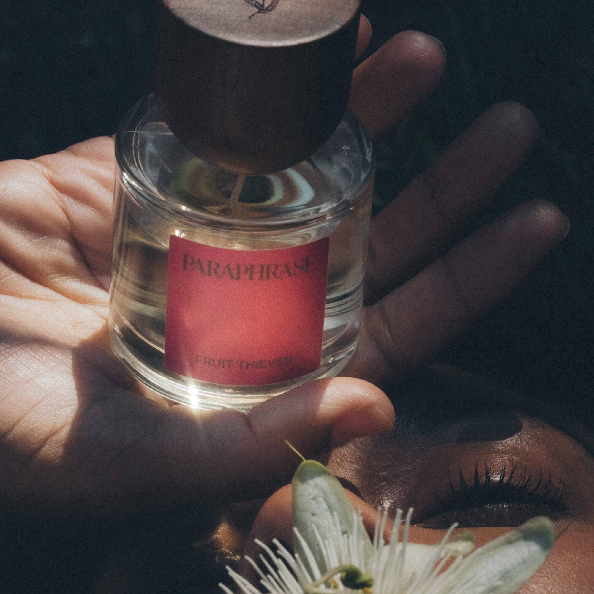 Fruit Thieves fragrance bottle near white flower and closed eye with sunlight highlights