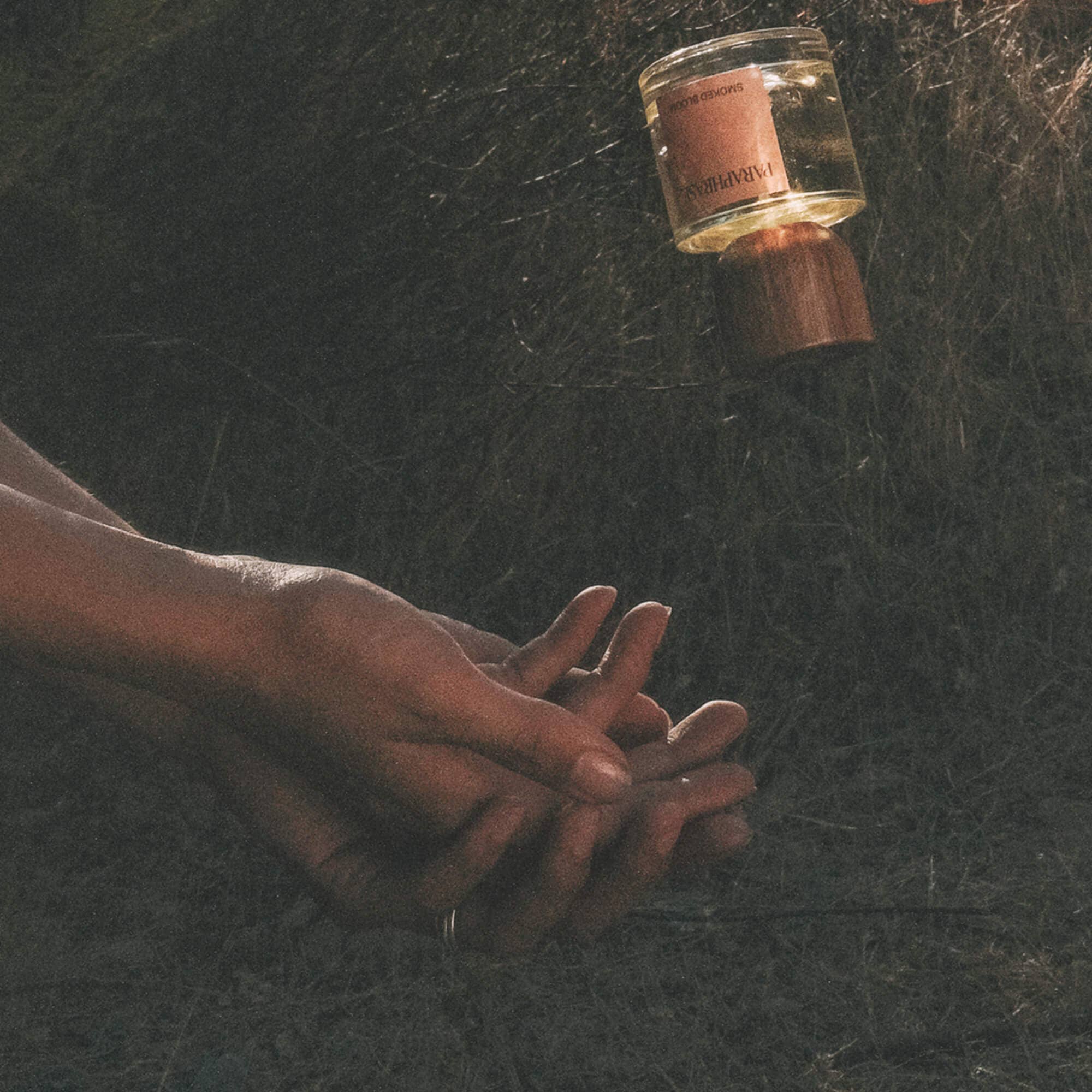 Floating Paraphrase Smoked Bloom perfume above hands in grass